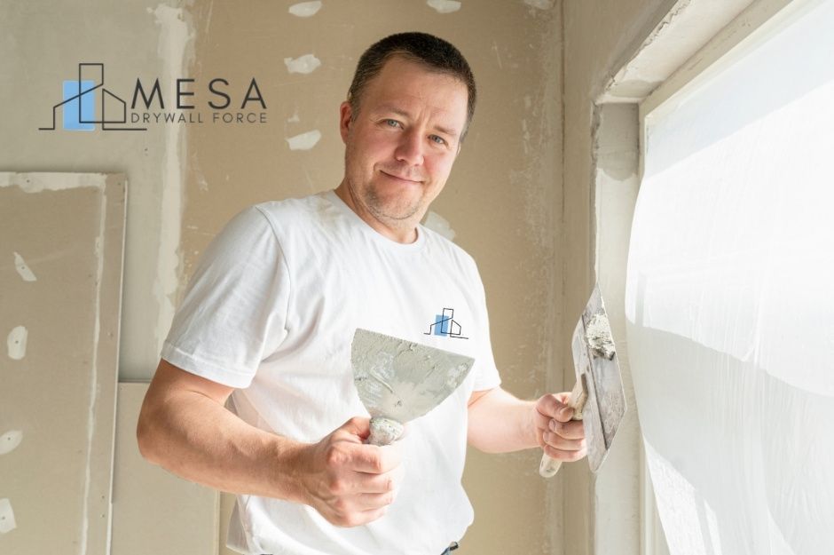 A drywall repair handyman applies joint compound using a putty knife during a residential project. He wears a white shirt and smiles while working on the drywall seams at a residential construction site, with patch repairs near W Nova Pl, and N Thunderbird Ave, Casa Grande, AZ