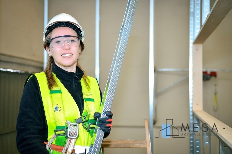 A drywall repair handyman in a yellow safety vest and white hard hat smiles at a commercial construction site with exposed wall framing. She wears safety glasses and stands near partially completed interior walls with metal studs visible near S 204th Dr, and S 203rd Dr, Liberty, AZ.
