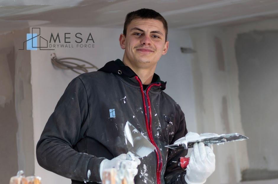 A drywall repair handyman at a residential construction site holding a putty knife and trowel, wearing work gloves and a dark hoodie. White drywall compound is visible on the wall behind him near W Camino Del Sol, and N 137th Dr, Sun City West, AZ.