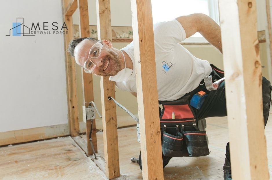 A drywall repair specialist wearing safety glasses, a white shirt, and a tool belt kneels between exposed wooden wall studs during a commercial renovation. He smiles while holding a pry bar and inspecting the wall framing. Located near N 64th Pl, and N 65th St, Paradise Valley, AZ.