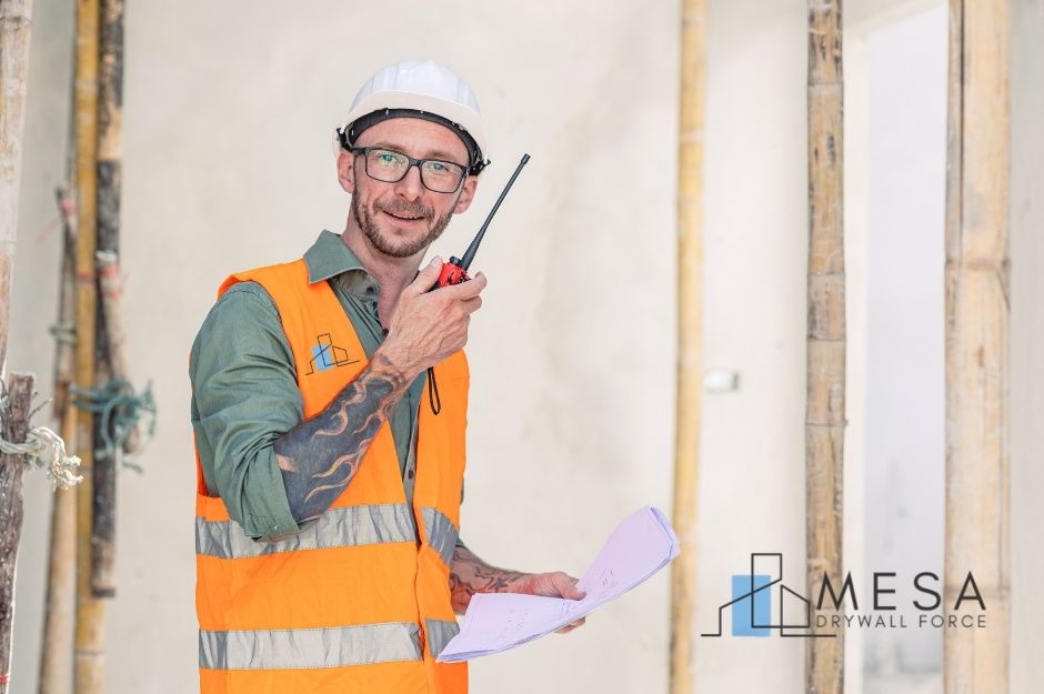 A drywall repair technician at a commercial construction site smiles while holding blueprints and a two-way radio. He wears a white hard hat, an orange high-visibility vest, and has tattoos on his arms. Wooden framing is visible in the background near W Cardinal Dr, and S 181st Ave, Goodyear, AZ.