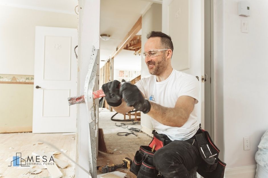 A drywall repair technician, wearing safety glasses and black gloves, uses a crowbar during a residential renovation project. He is dressed in a white shirt and a tool bag, working on drywall in a home under construction near E Buck Basin Dr, and N Green River Ln, Rio Verde, AZ.