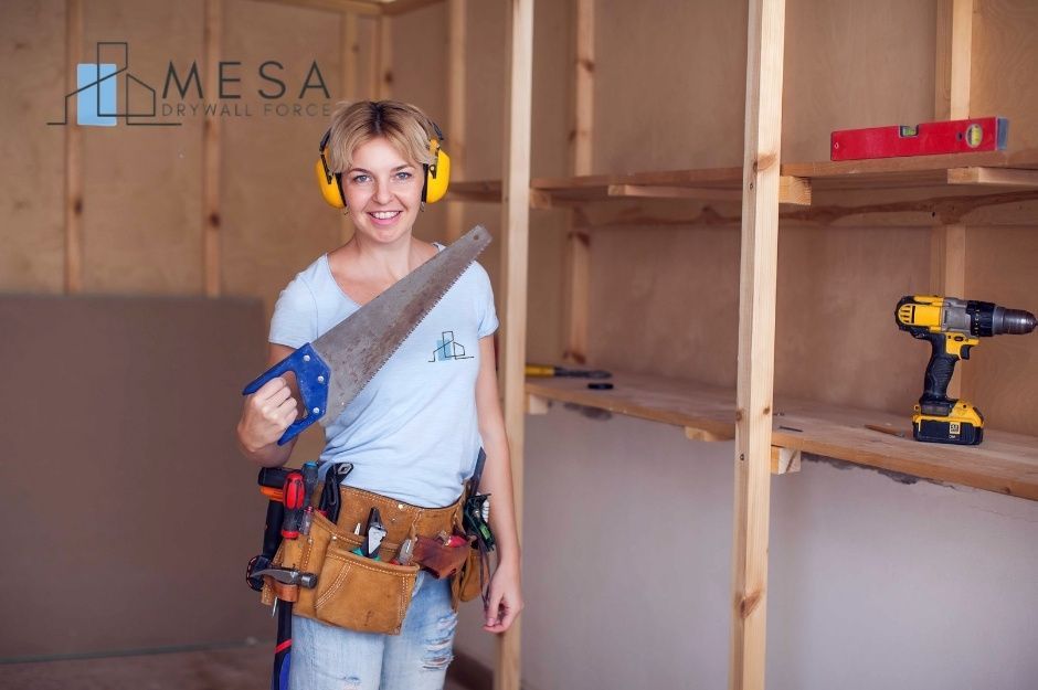 A drywall repair technician at a commercial construction site wearing yellow ear protection and a tool belt, holding a handsaw, smiling in an unfinished wood-framed room with a level and cordless drill visible on shelving, near N 42nd Ln, and W Acorn Valley Trail, Rio Vista, AZ.
