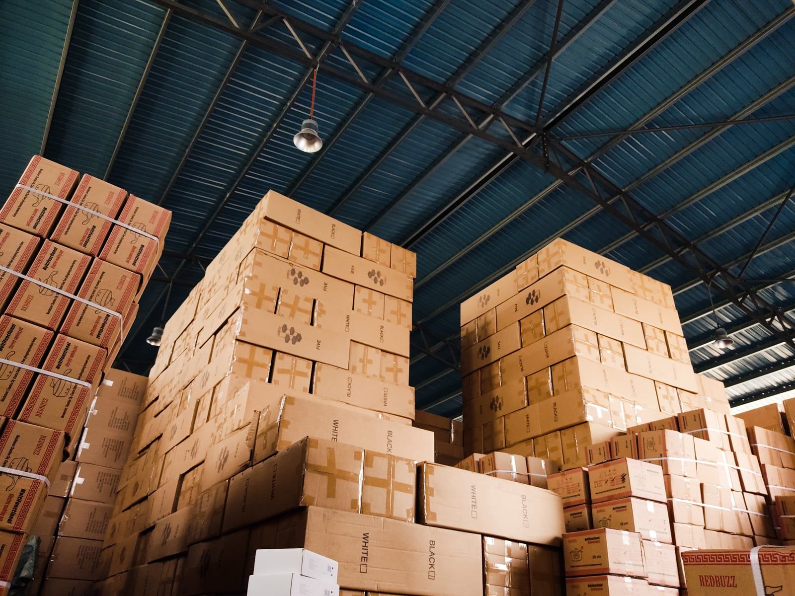 Stacks of cardboard boxes piled high in a warehouse with a blue metal ceiling