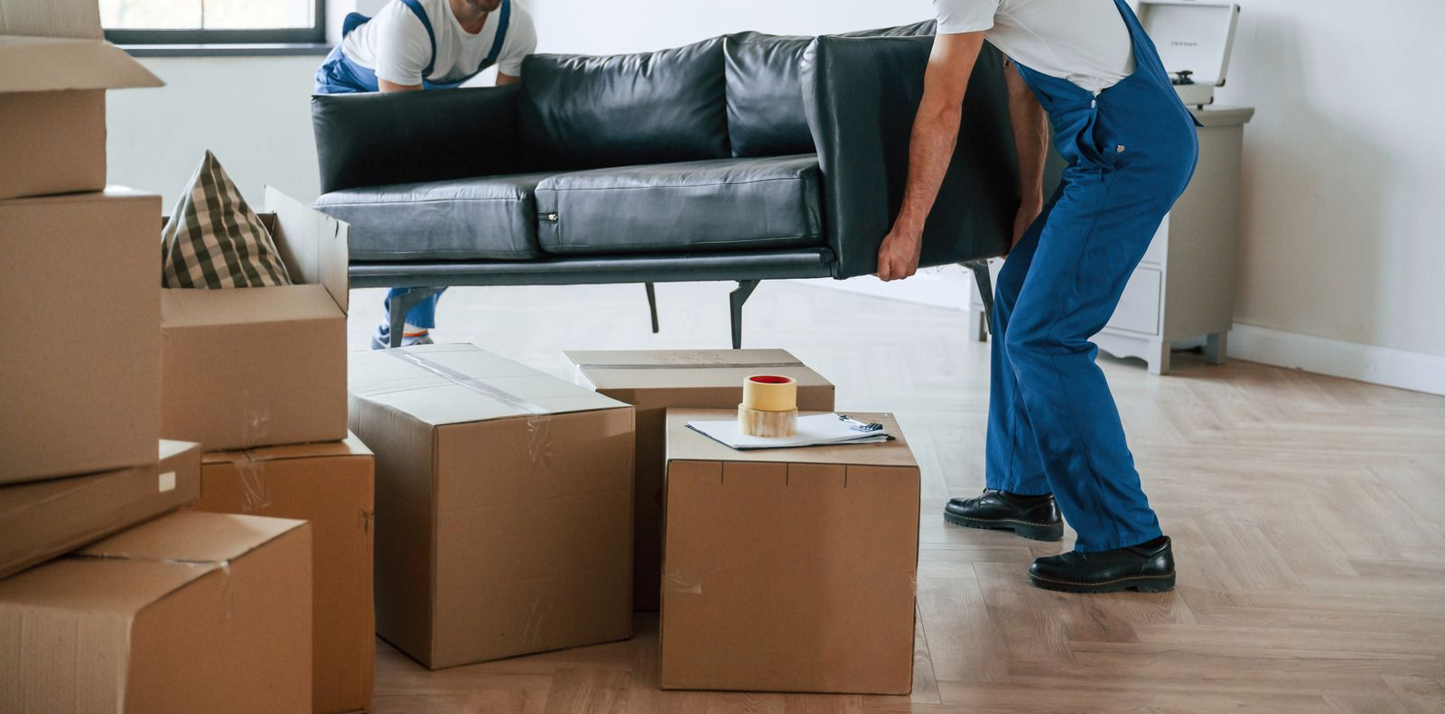 Movers carrying a black sofa through a living room with cardboard boxes stacked on the floor