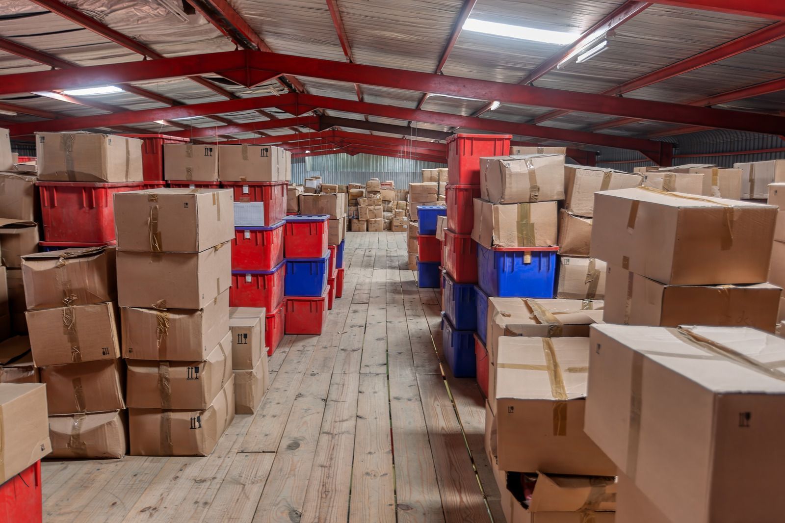 Warehouse aisle stacked with cardboard boxes and red-blue crates under a corrugated metal roof