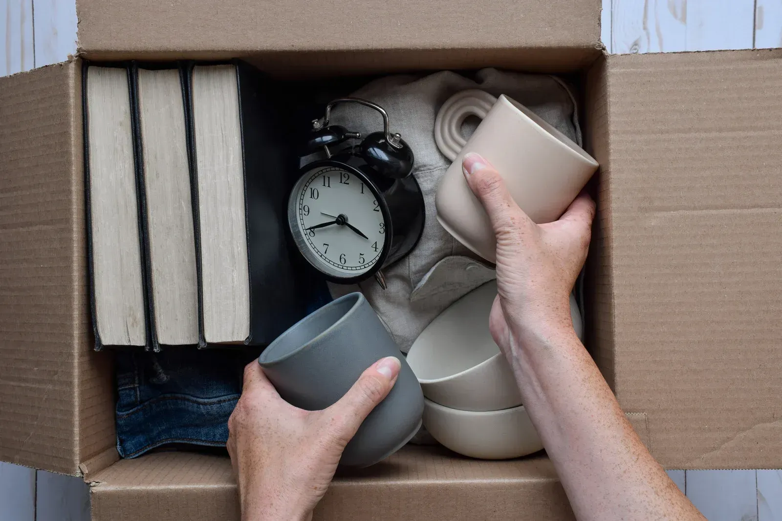 Hands packing mugs and an alarm clock into a cardboard box