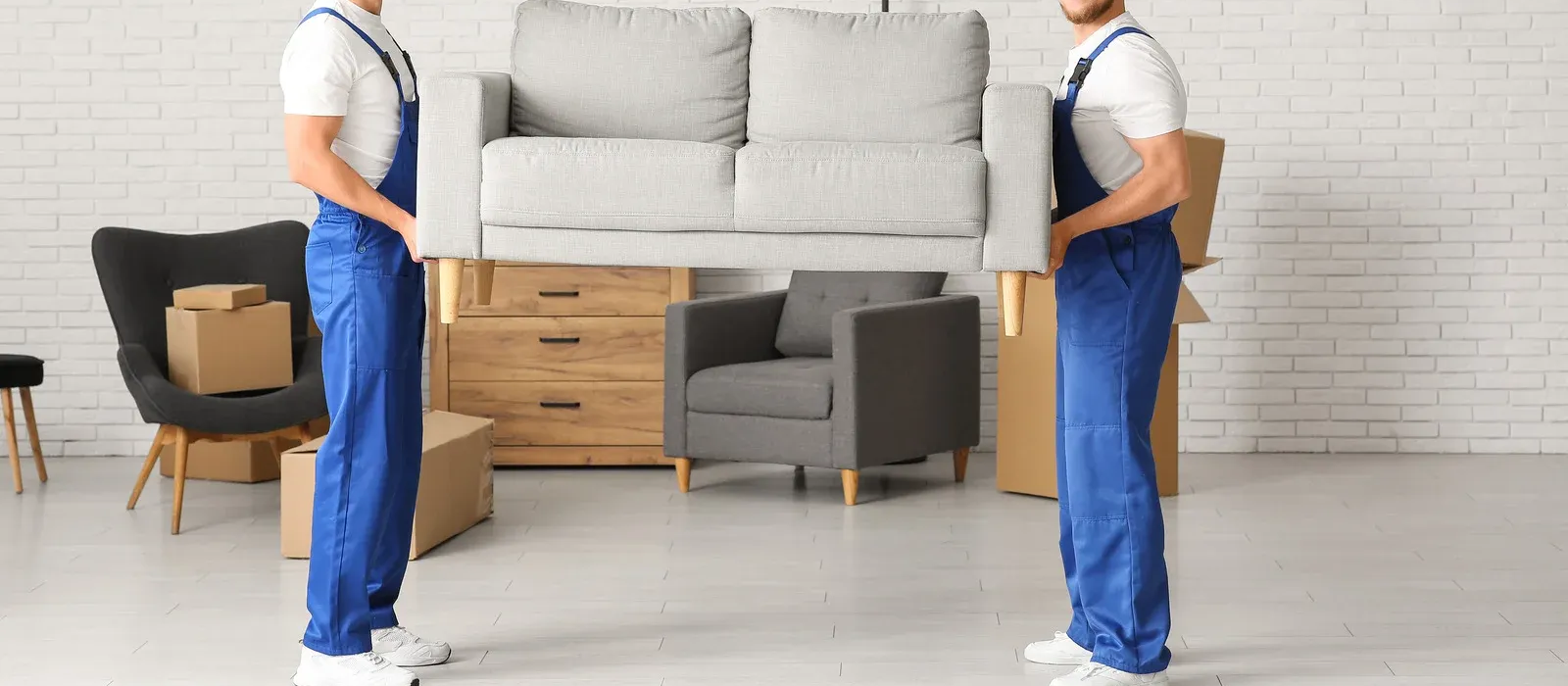 Two movers carrying a sofa into a living room with stacked furniture and boxes