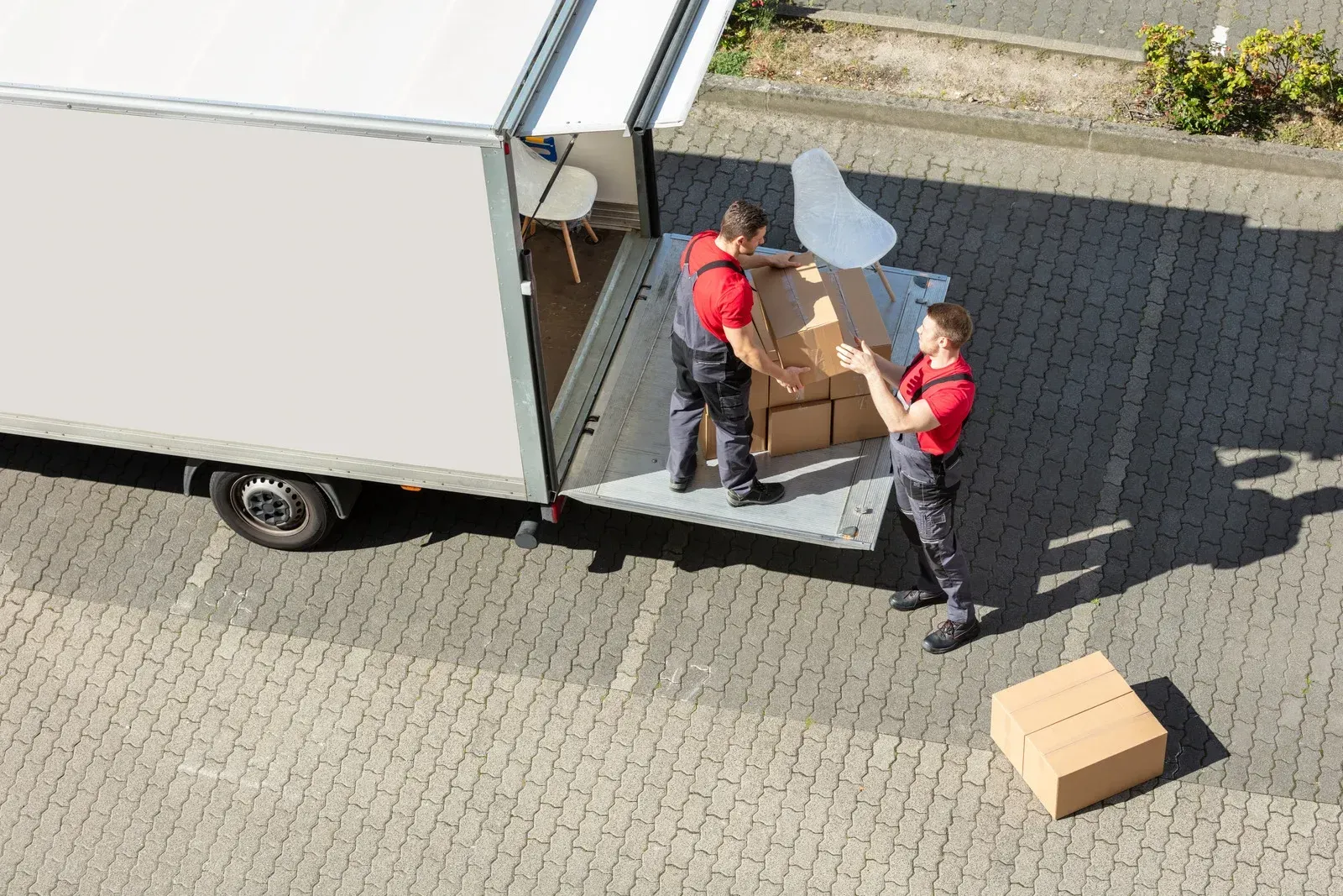 Two workers in red shirts unload boxes from a delivery truck onto a paved lot.