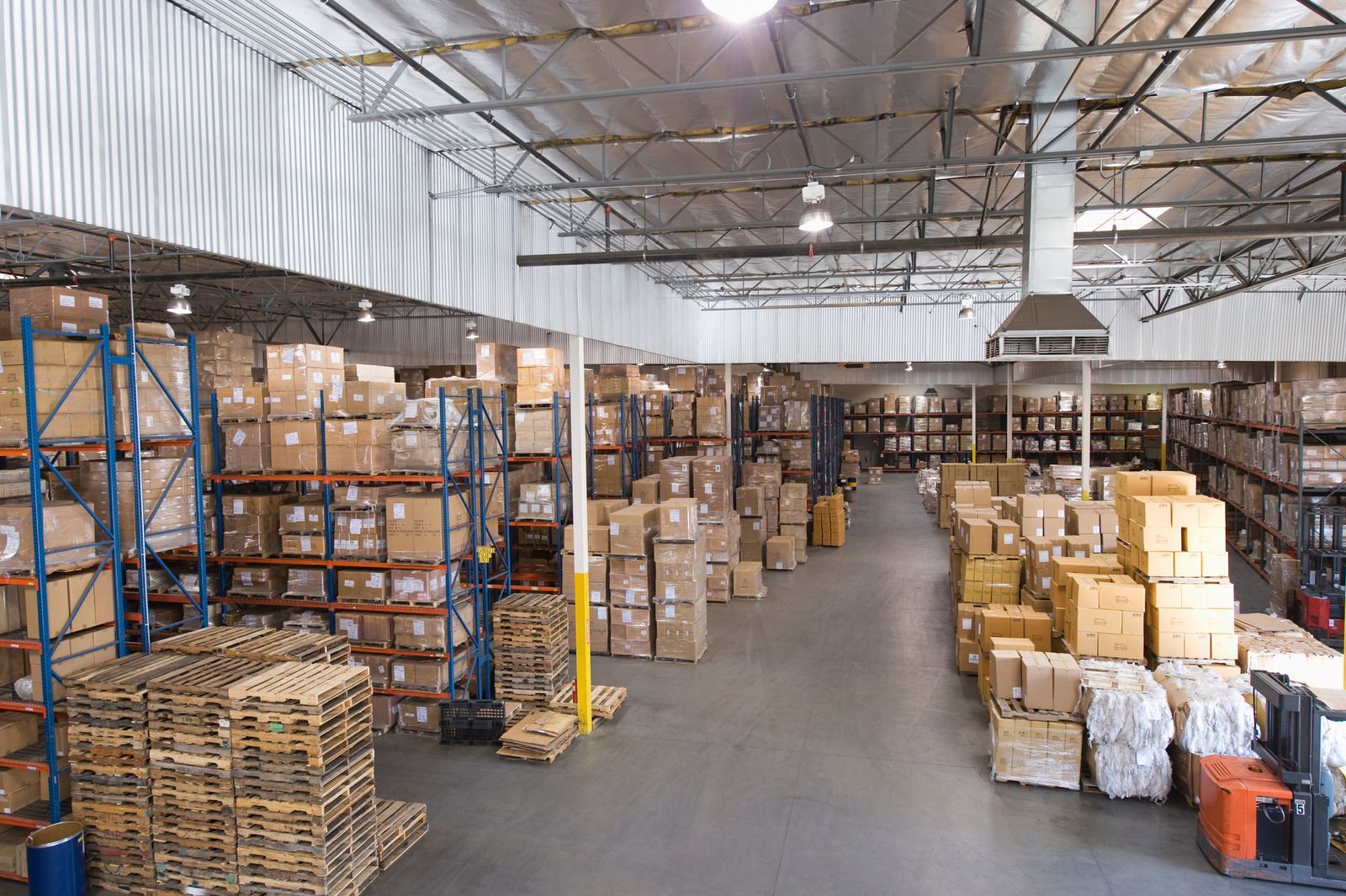 Warehouse aisle with stacked cardboard boxes, pallets, and metal shelving under a high ceiling