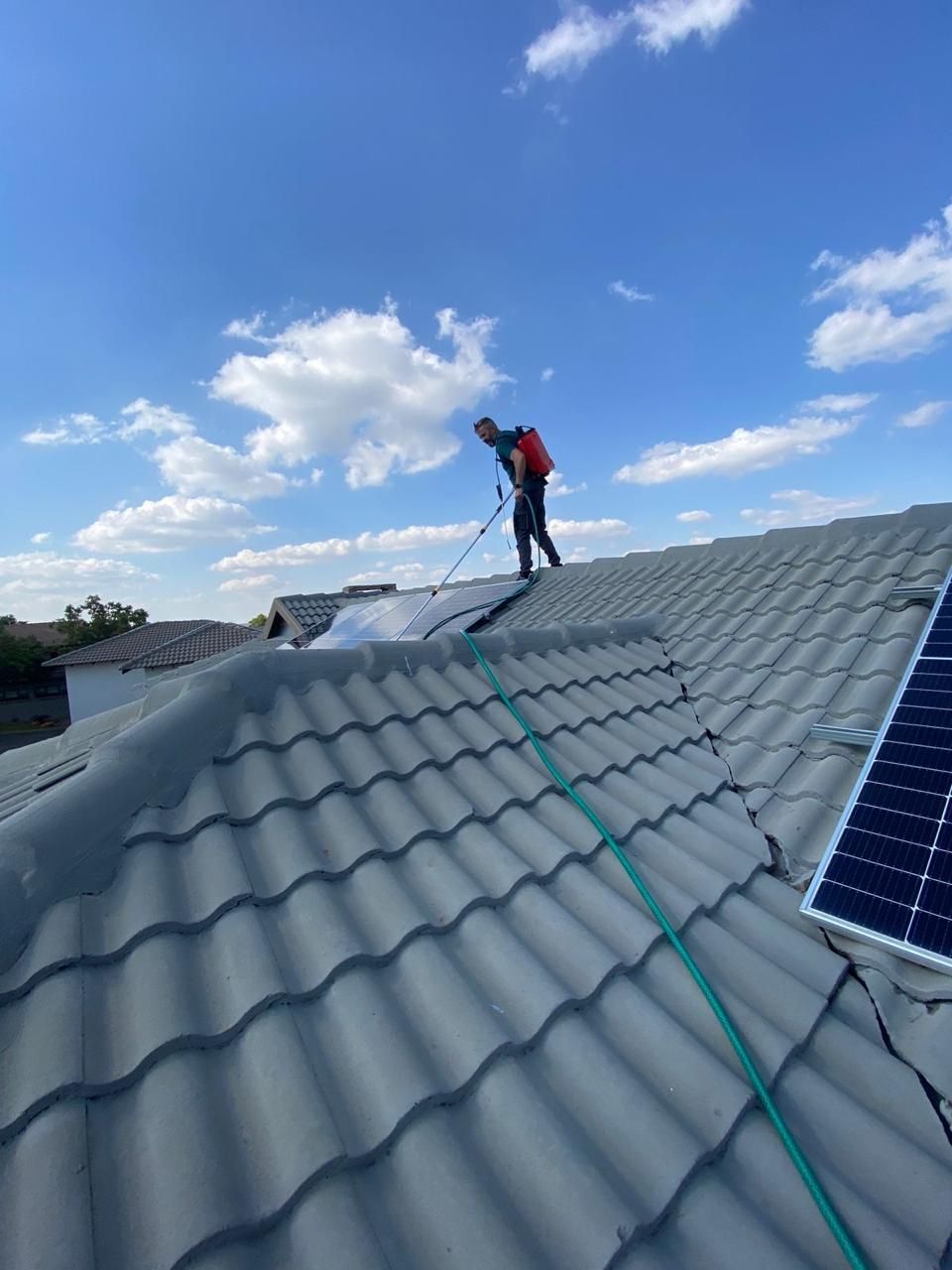 A worker in a red backpack sprayer cleans a tiled roof under a bright blue sky with scattered white clouds.