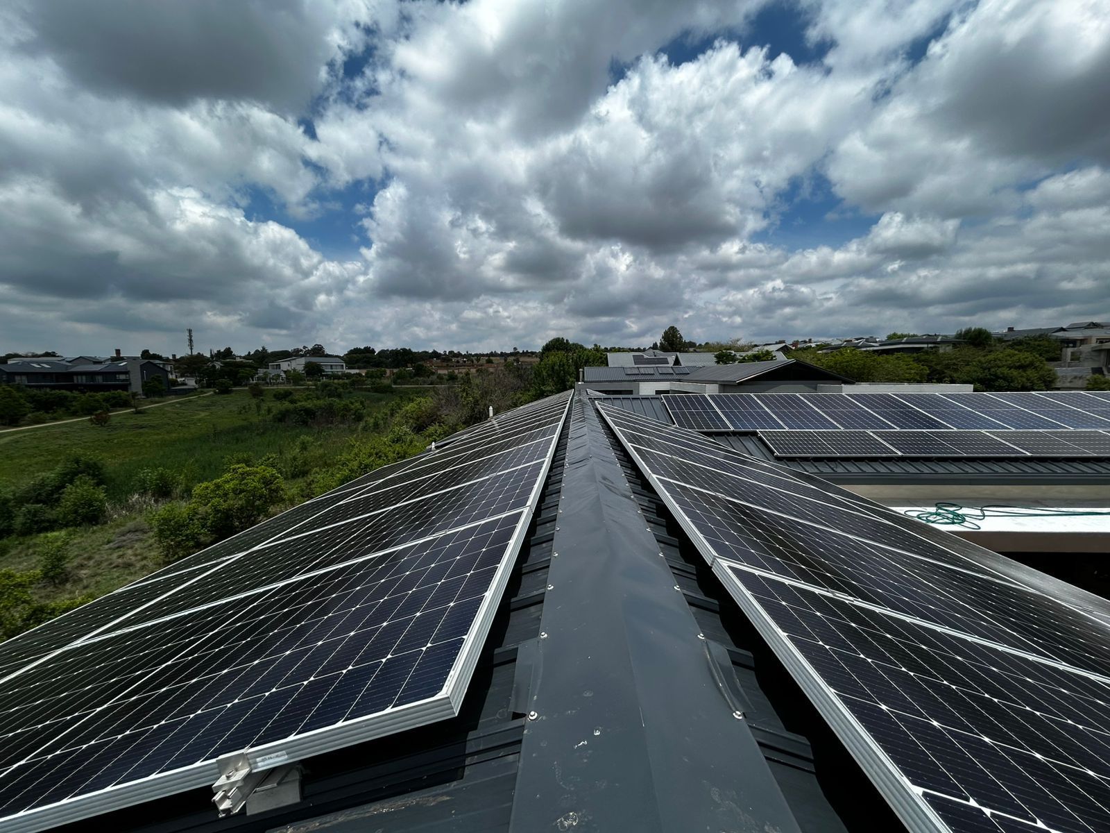Rows of dark solar panels mounted on a residential roof under a bright, cloudy sky overlooking a field.