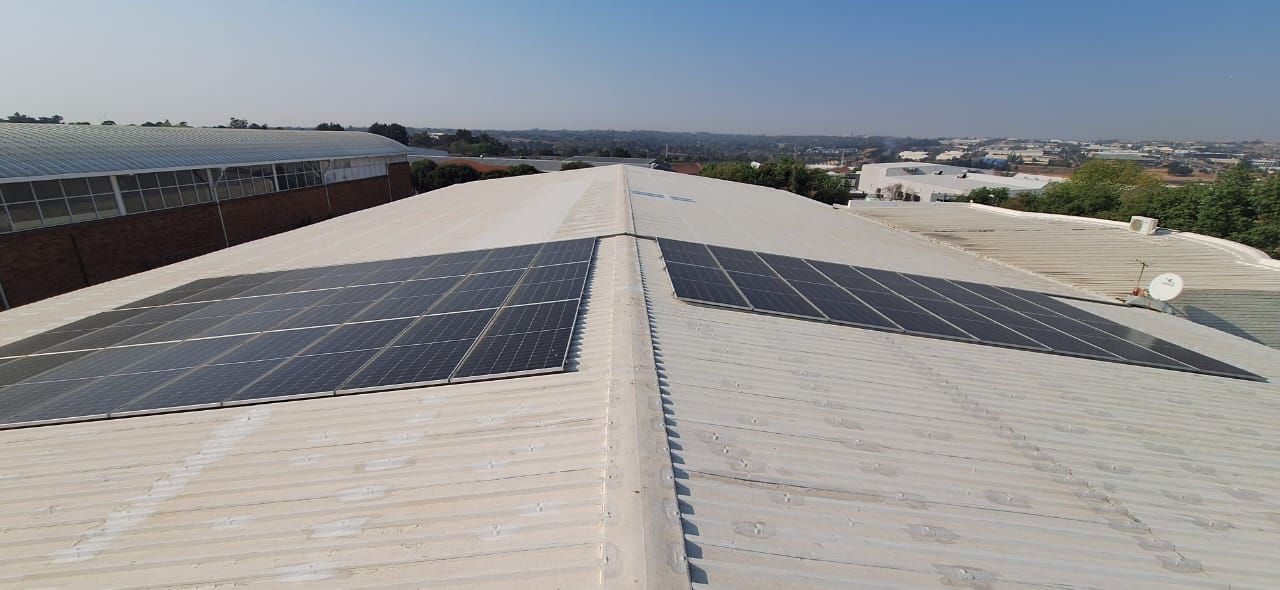 Two rectangular arrays of dark solar panels installed on a white industrial metal roof under a clear blue sky.