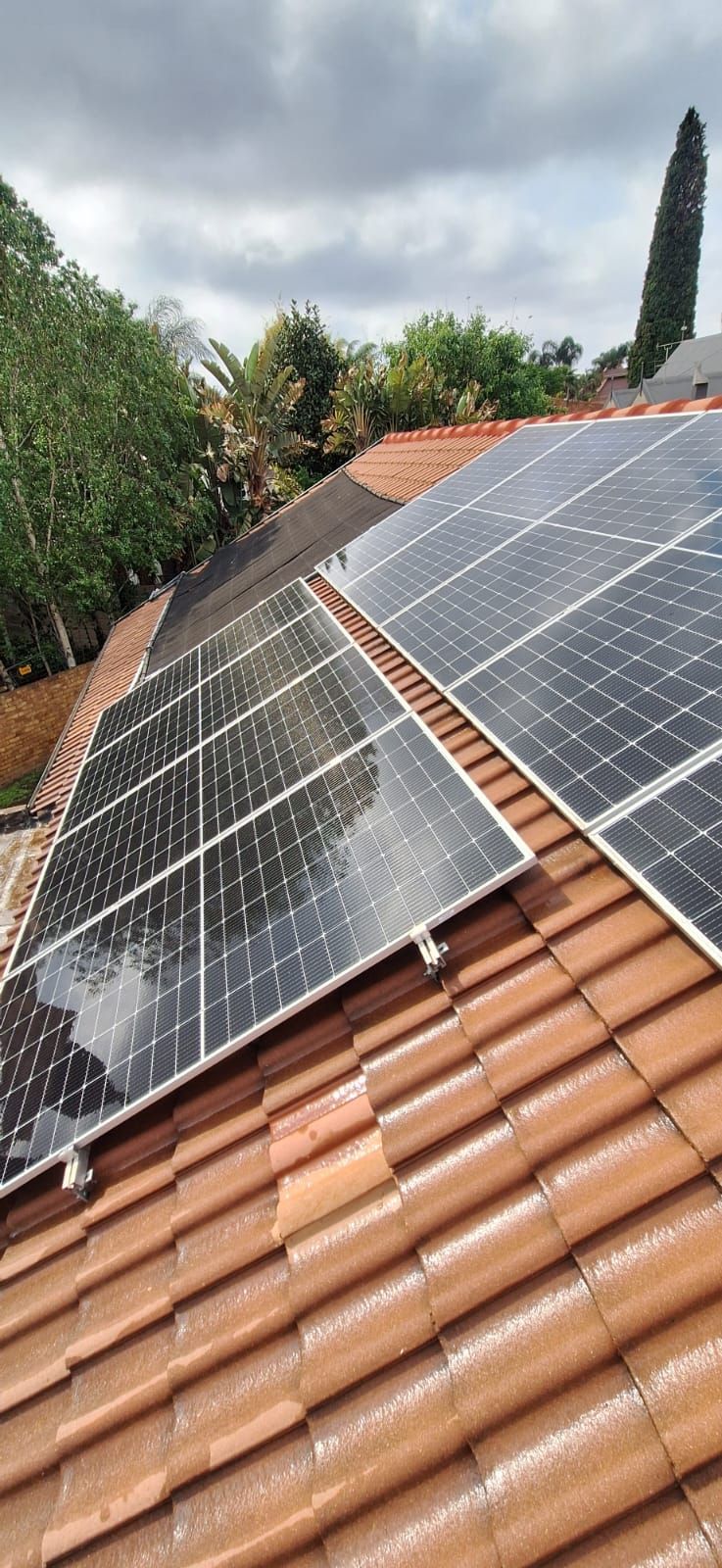 Solar panels installed on a residential terracotta tiled roof under a cloudy sky with trees in the background.