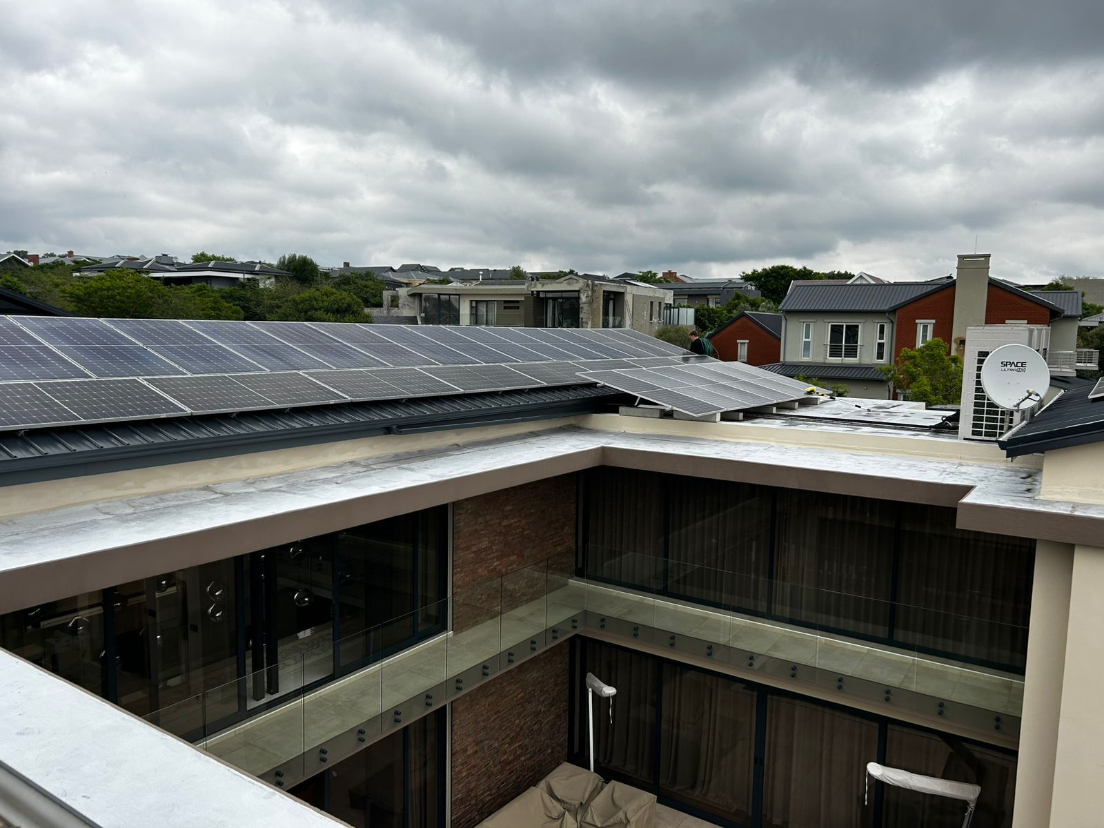 Solar panels installed on a residential roof above a two-story building with a courtyard under a cloudy sky.