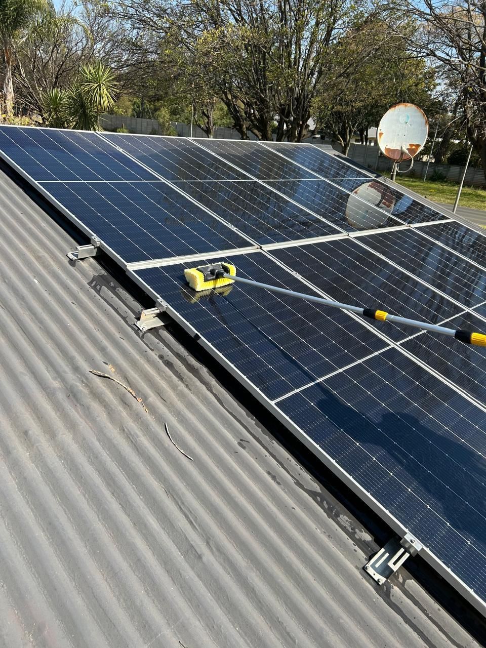 A person uses a long-handled cleaning brush to scrub solar panels installed on a corrugated metal roof.