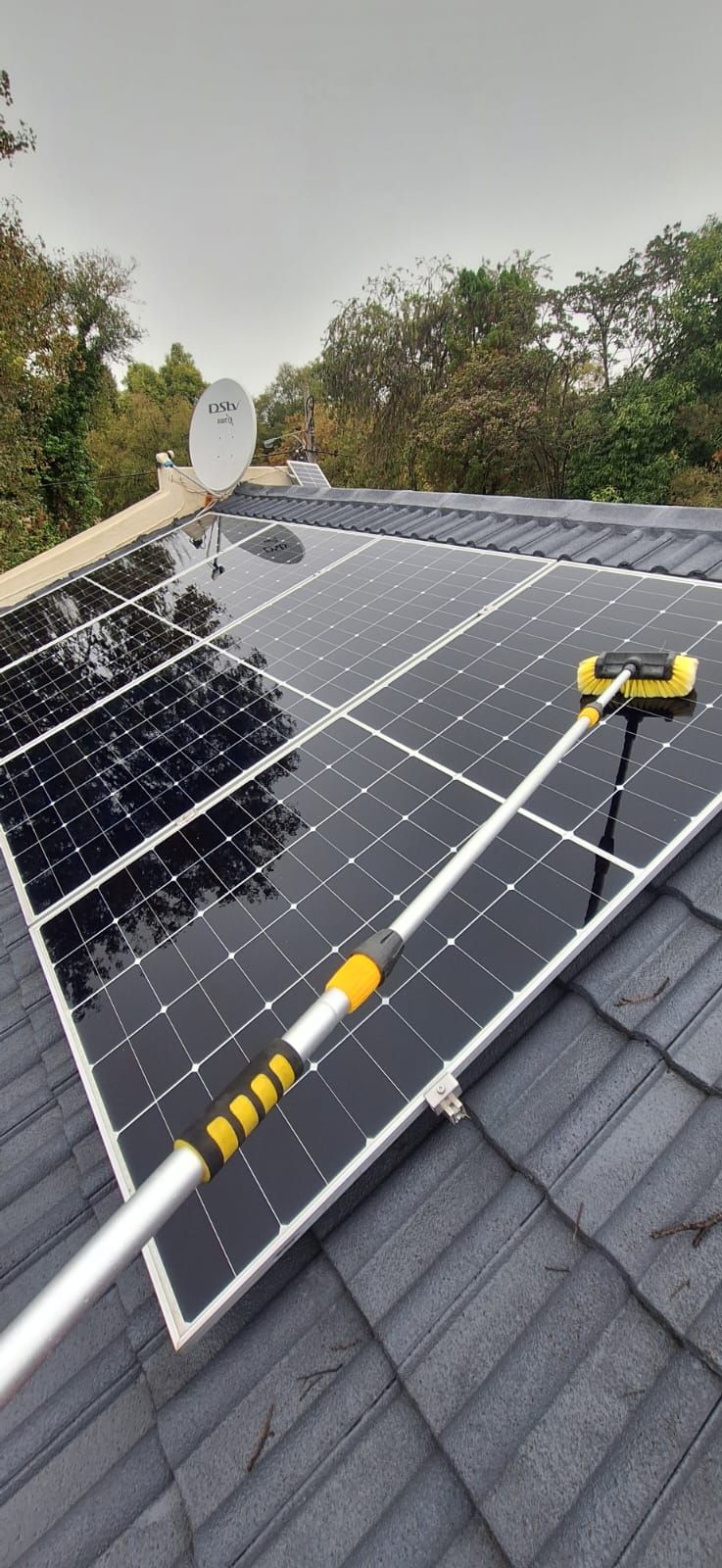 A telescopic pole with a brush cleaning solar panels installed on a dark tiled roof, with trees in the background.
