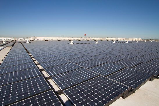 An expansive array of dark blue solar panels installed across a flat, light-colored industrial rooftop under a blue sky.