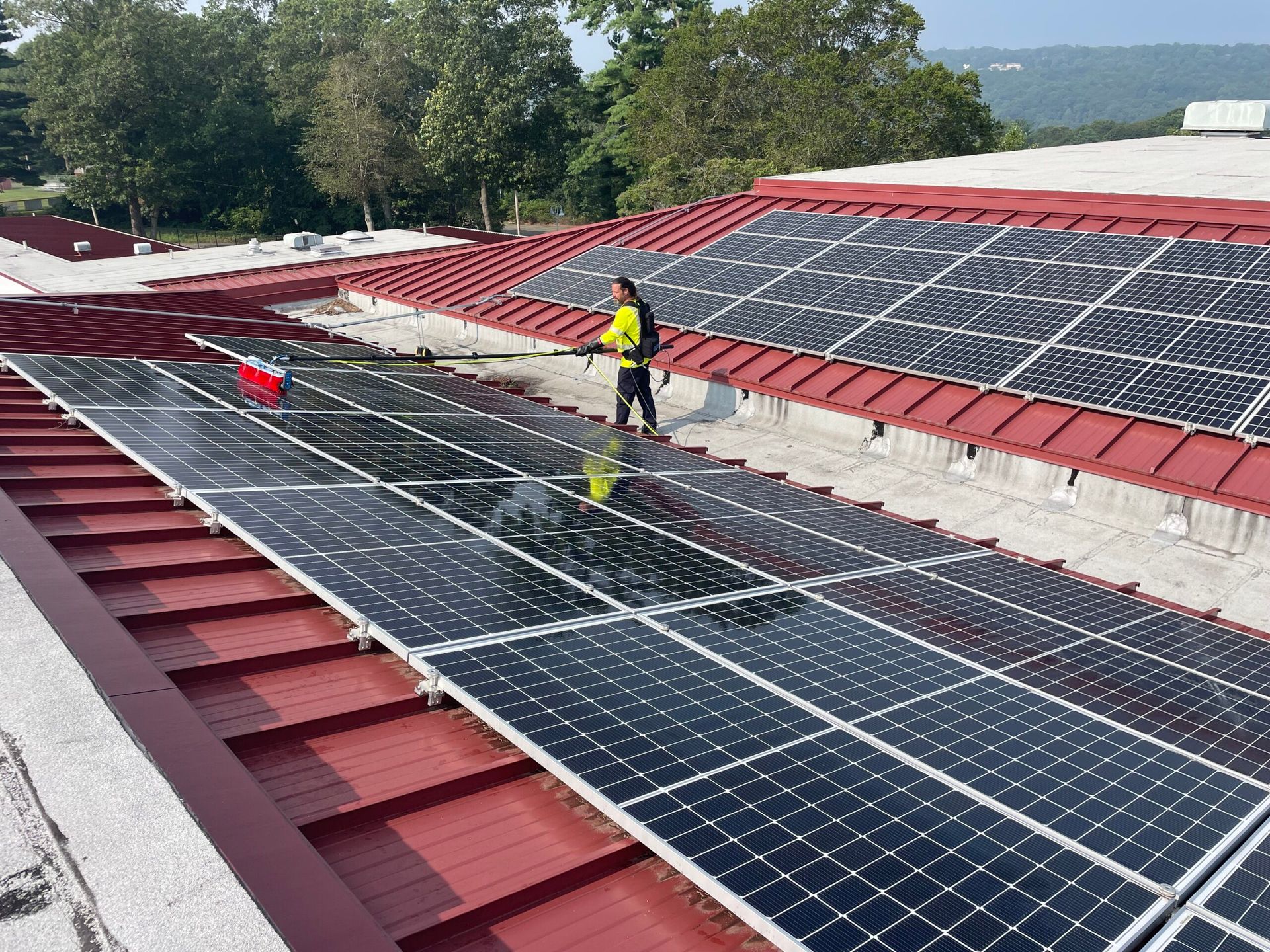 A person in a high-visibility vest cleans solar panels on a red metal roof using a long-handled brush.