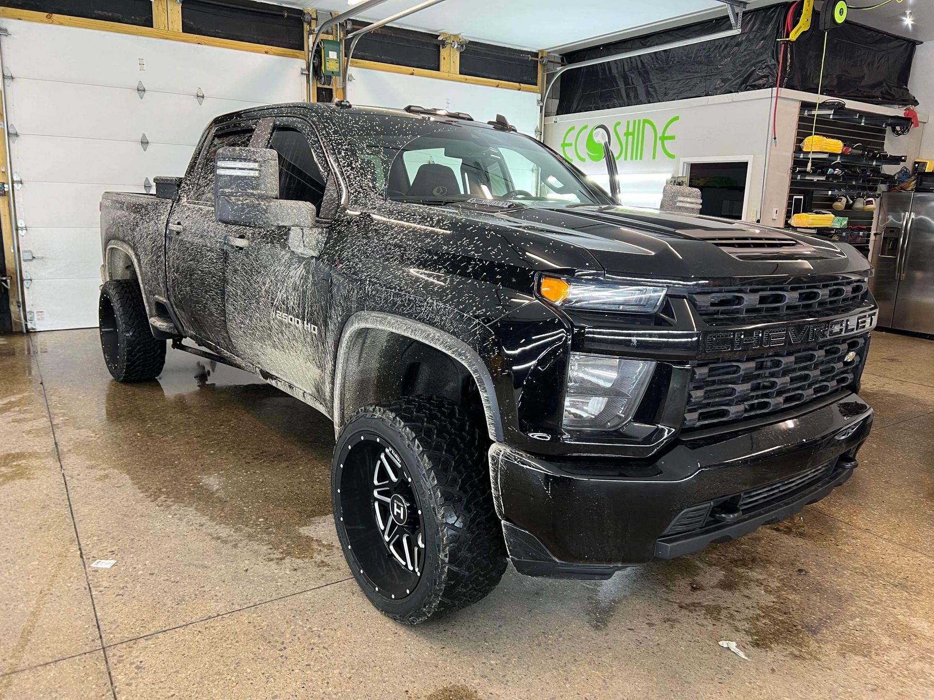 Black lifted pickup truck in a garage, covered in mud and snow, viewed from the front-left.