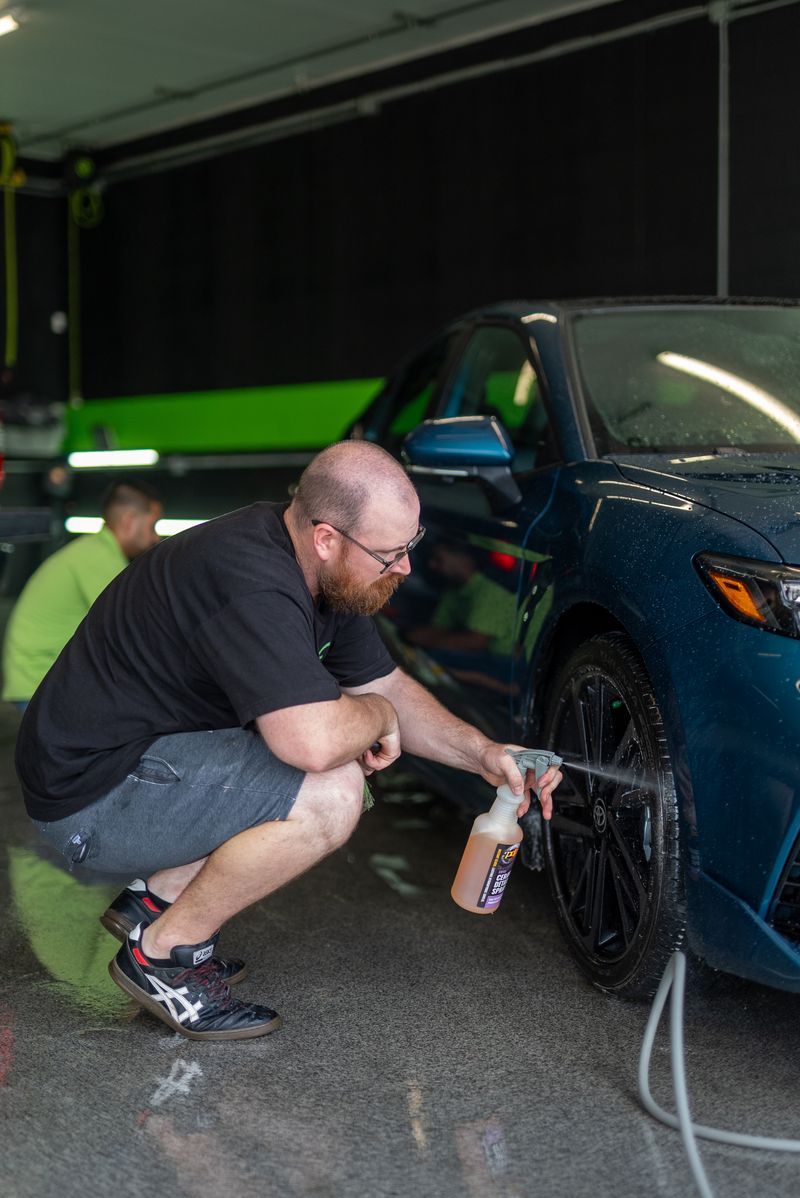 Man crouching to clean a blue car wheel in a garage, holding a spray bottle.