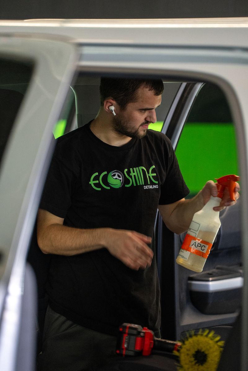 Man holding a spray bottle and tool beside a van, wearing a black T-shirt in a garage setting
