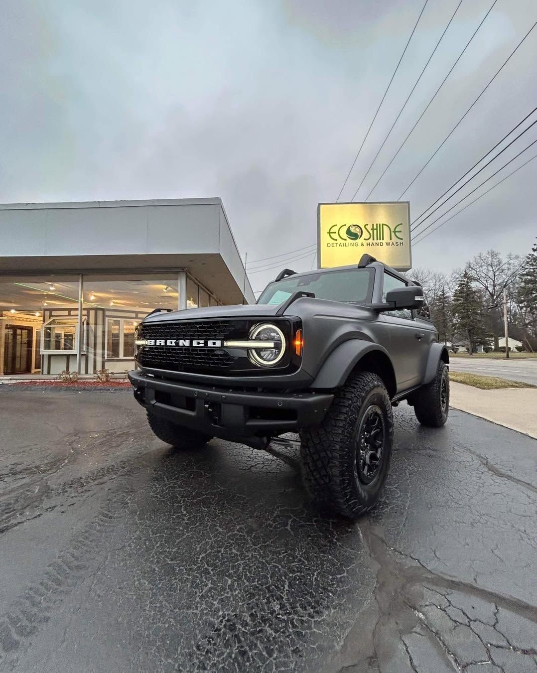 Black off-road truck parked outside a store on a wet street under a cloudy sky.