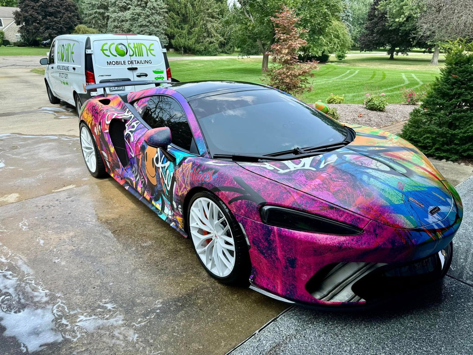 Colorfully wrapped sports car parked on a driveway with a white work van behind it.
