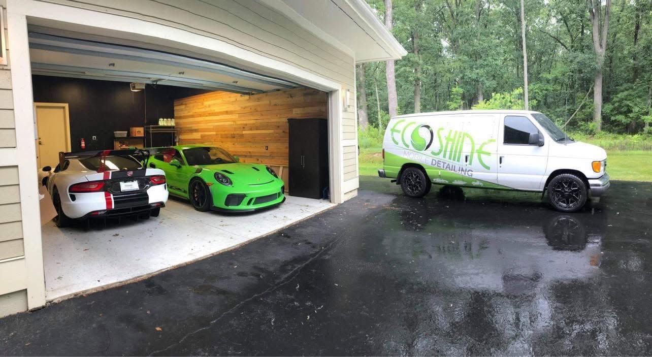 Two sports cars in a garage and a white service van parked on a wet driveway