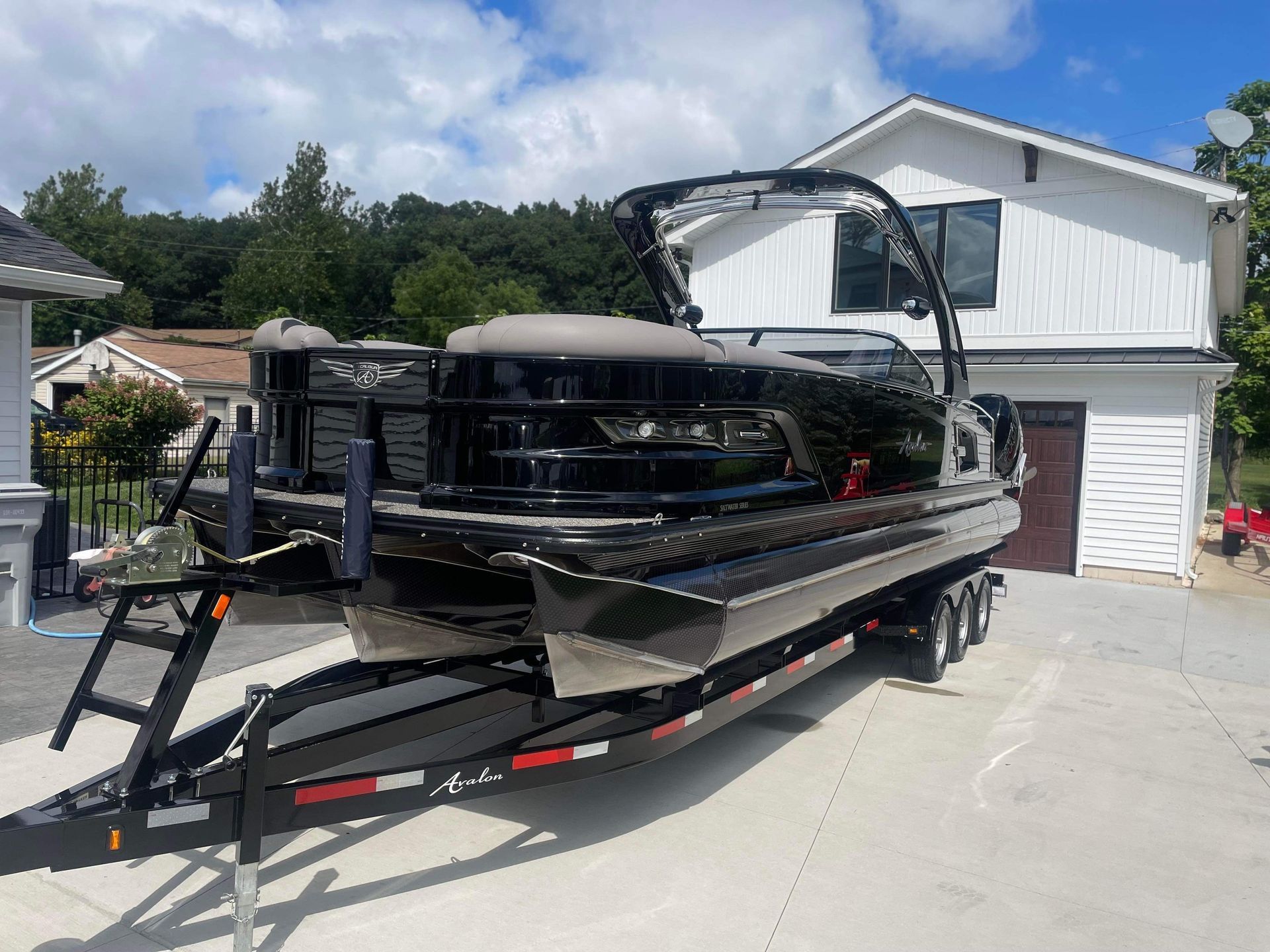 Black pontoon boat on a trailer in a driveway, with a basketball hoop and white house in the background