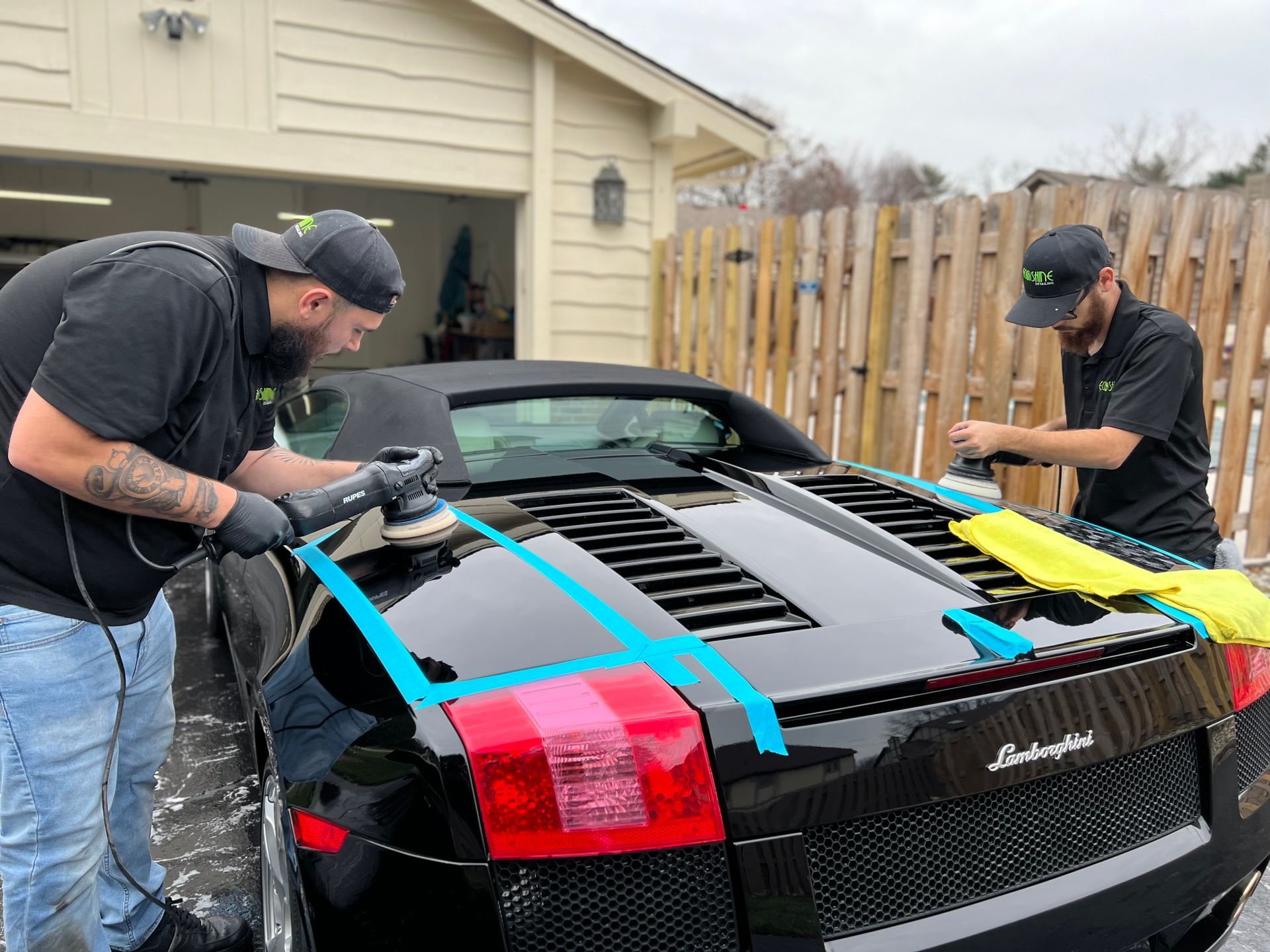 Two people taping blue painter’s tape on a black Lamborghini in a driveway