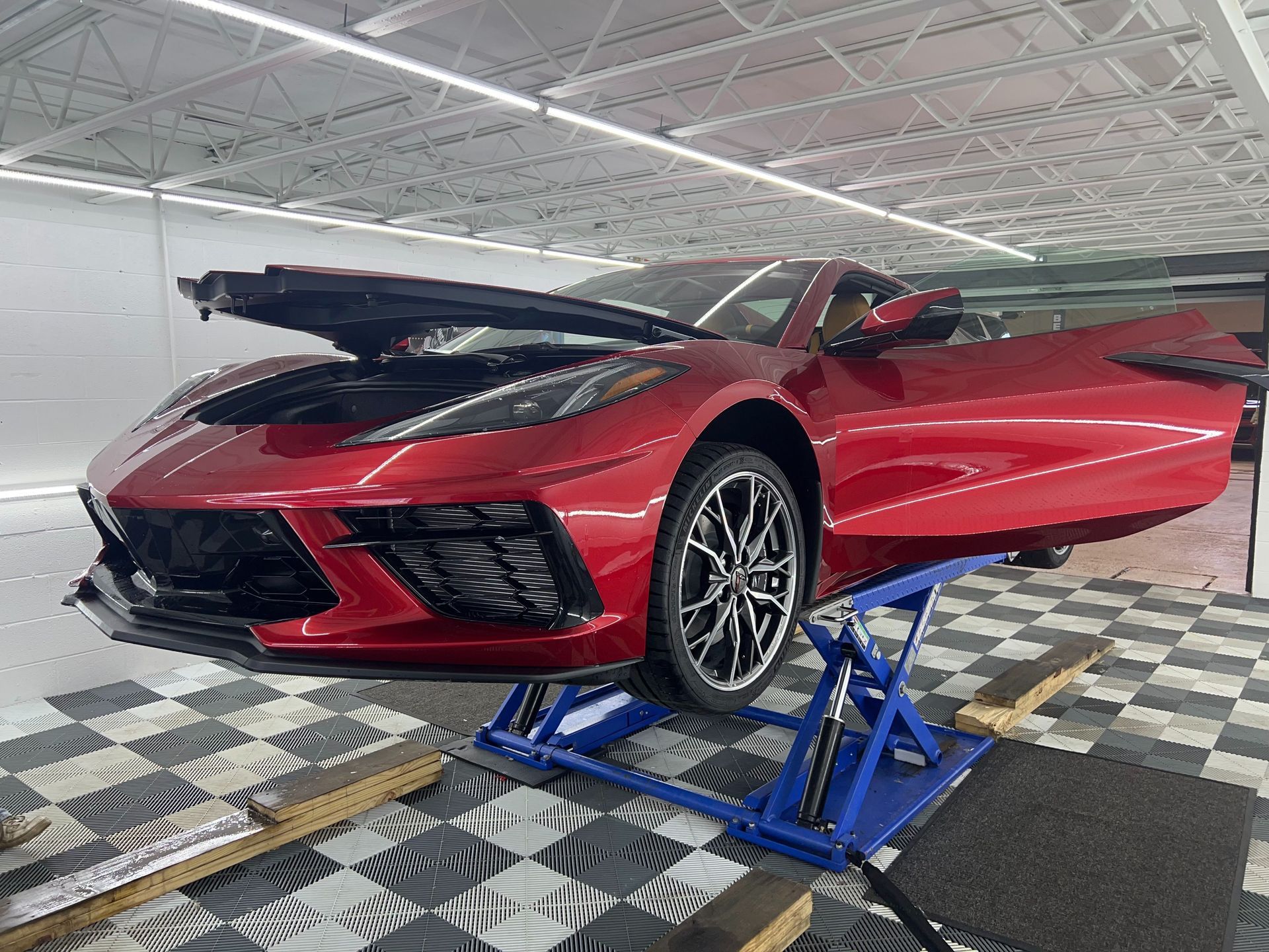 Red sports car raised on blue lift in a garage, with front hood open and driver door ajar.