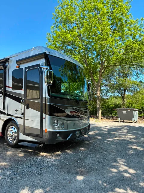 Silver RV parked on a gravel lot under trees on a sunny day