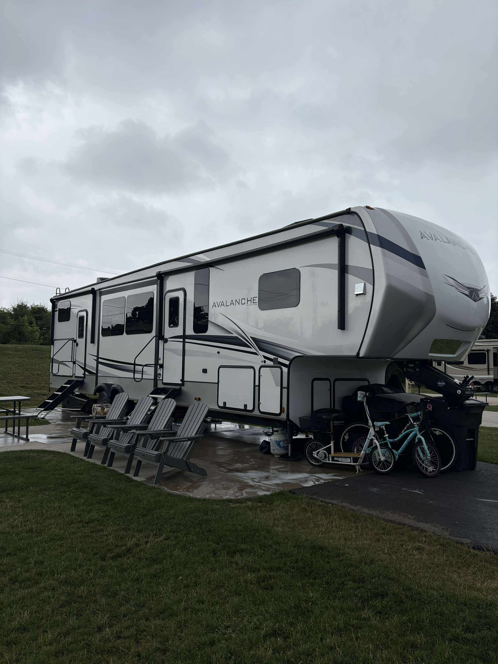 Large gray RV trailer parked on wet grass under cloudy skies, with folding chairs and bicycles nearby