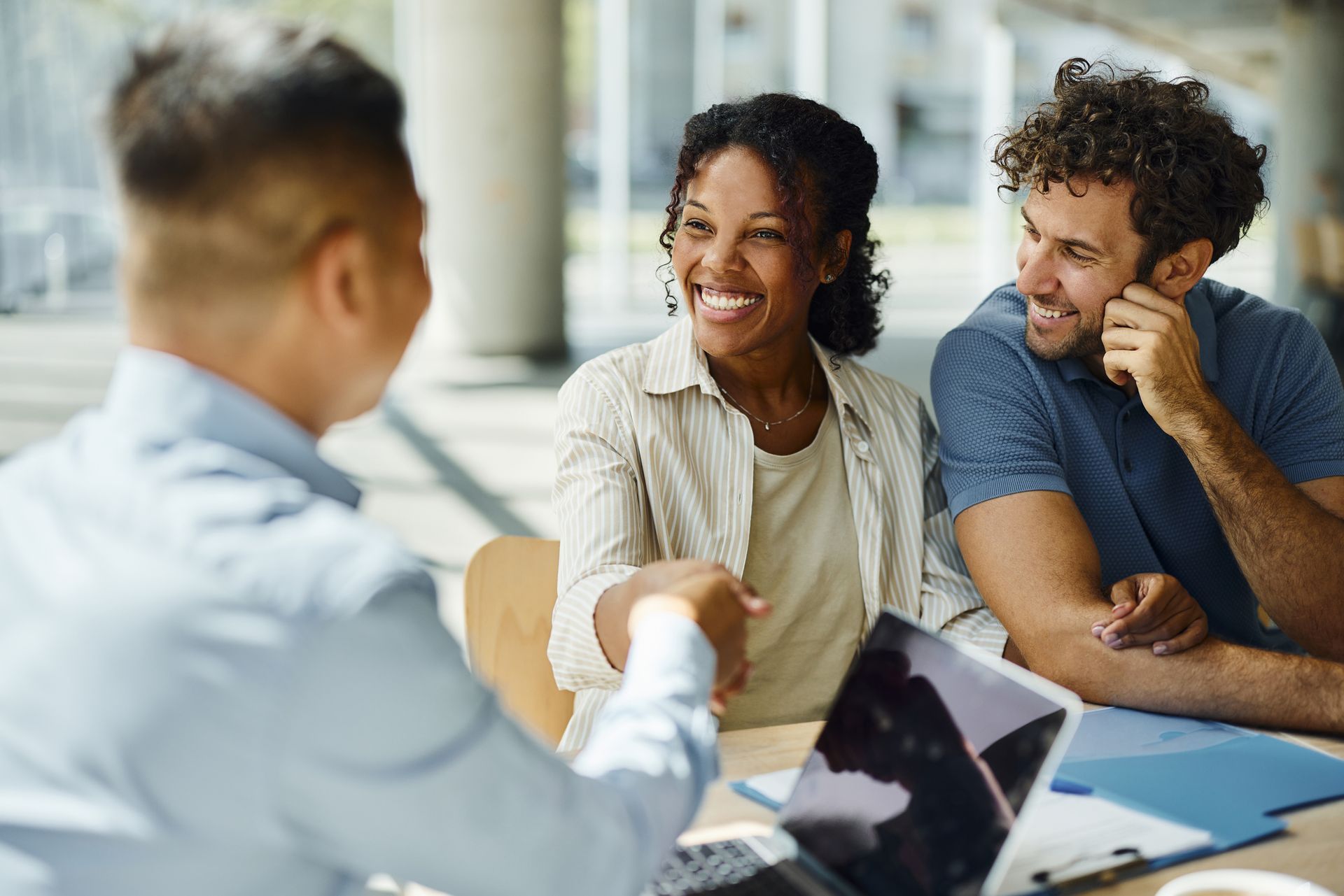Man shakes hands with a smiling woman while a man smiles. Meeting takes place in an office with a laptop on the table.