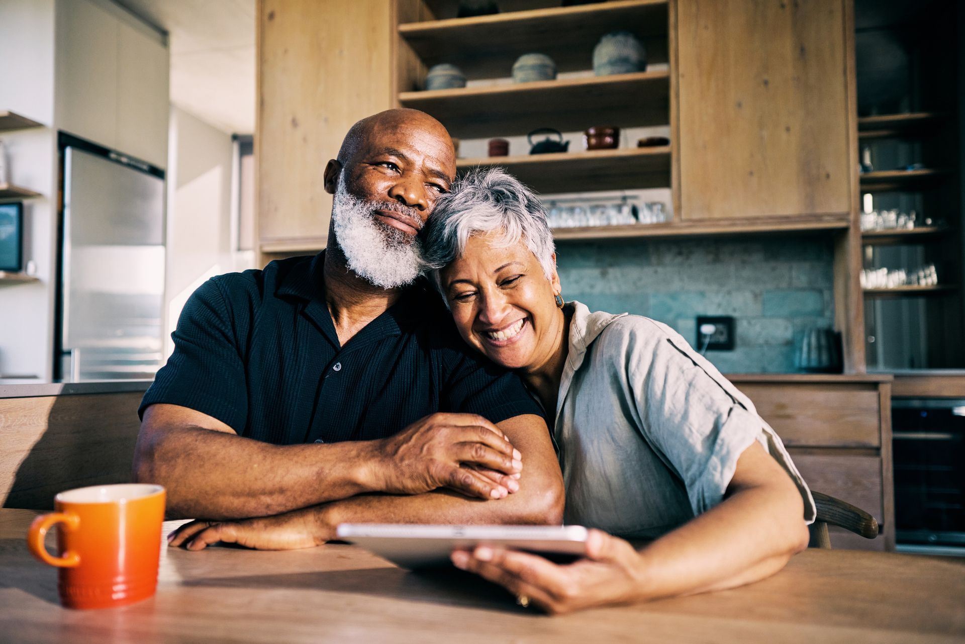 Smiling couple looks at tablet in kitchen. One person has head on other's shoulder. Orange mug on table.