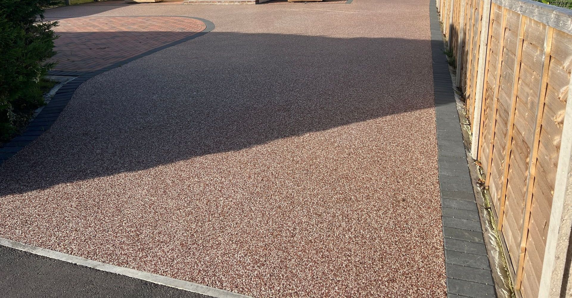 Gravel driveway, pinkish-brown stones, bordered by a dark edge and wooden fence.