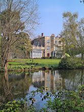 A large house is reflected in the water of a lake surrounded by trees.