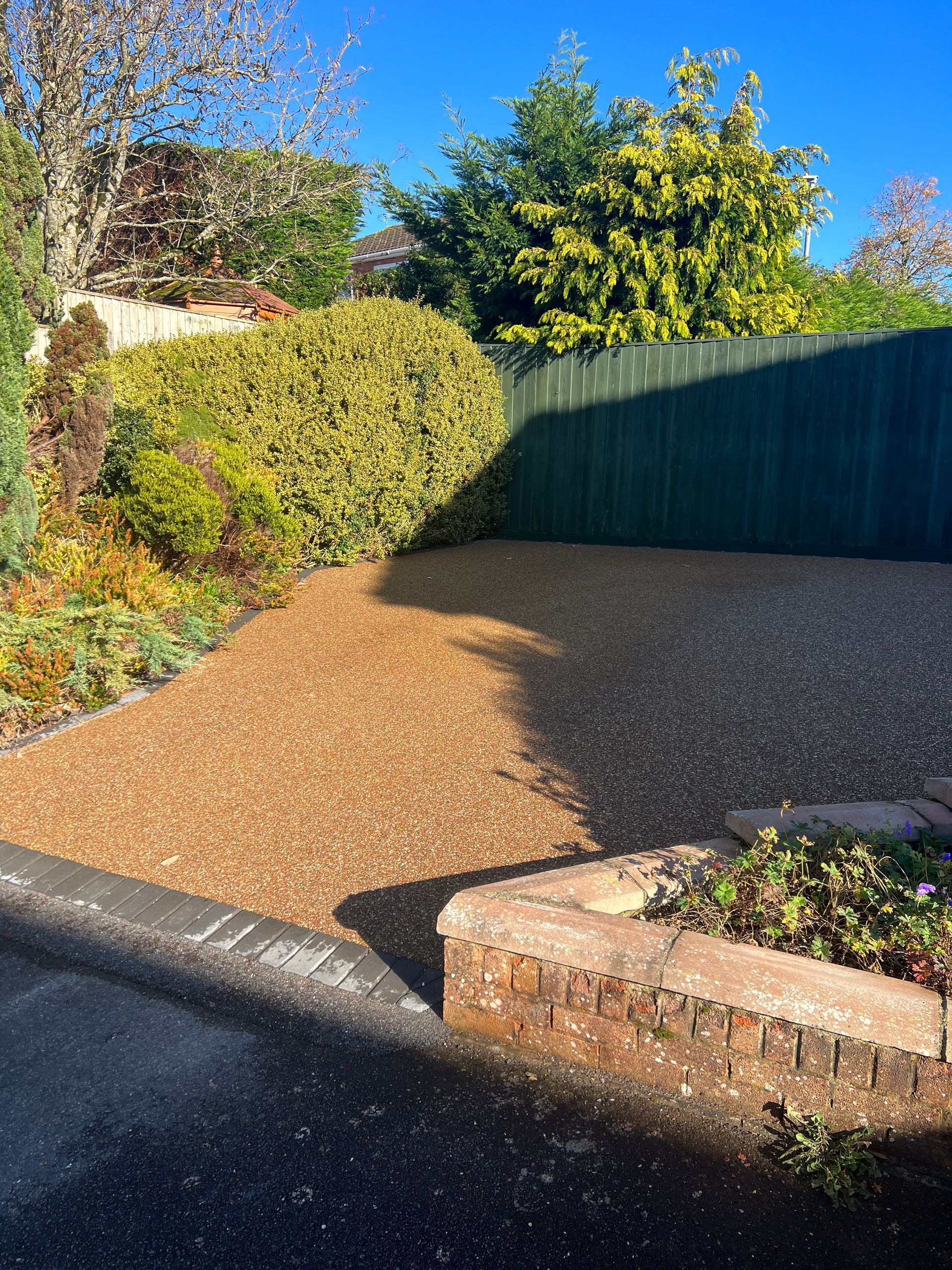Gravel driveway with a brick border, bushes, and a wooden fence under a blue sky.
