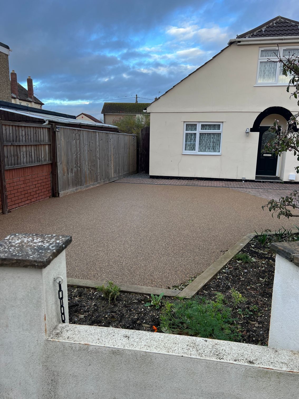 Beige house with gravel driveway, low wall with numbers in foreground, wooden fence on the left.