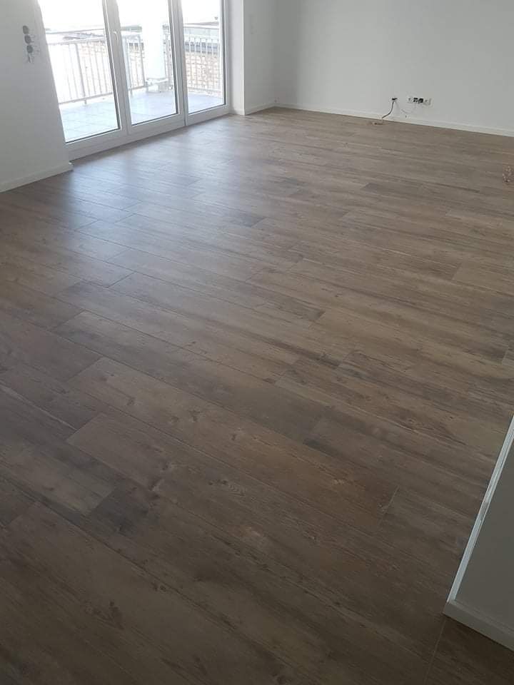 Leeres Zimmer mit Holzbodenoptik. Im Hintergrund befinden sich große Glastüren zu einem Balkon. Weiße Wände.