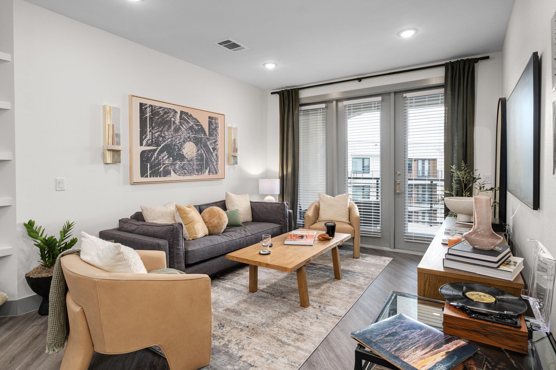 A modern living room with a gray sofa, two tan armchairs, and a wood coffee table. Large glass balcony doors, stylish rug.