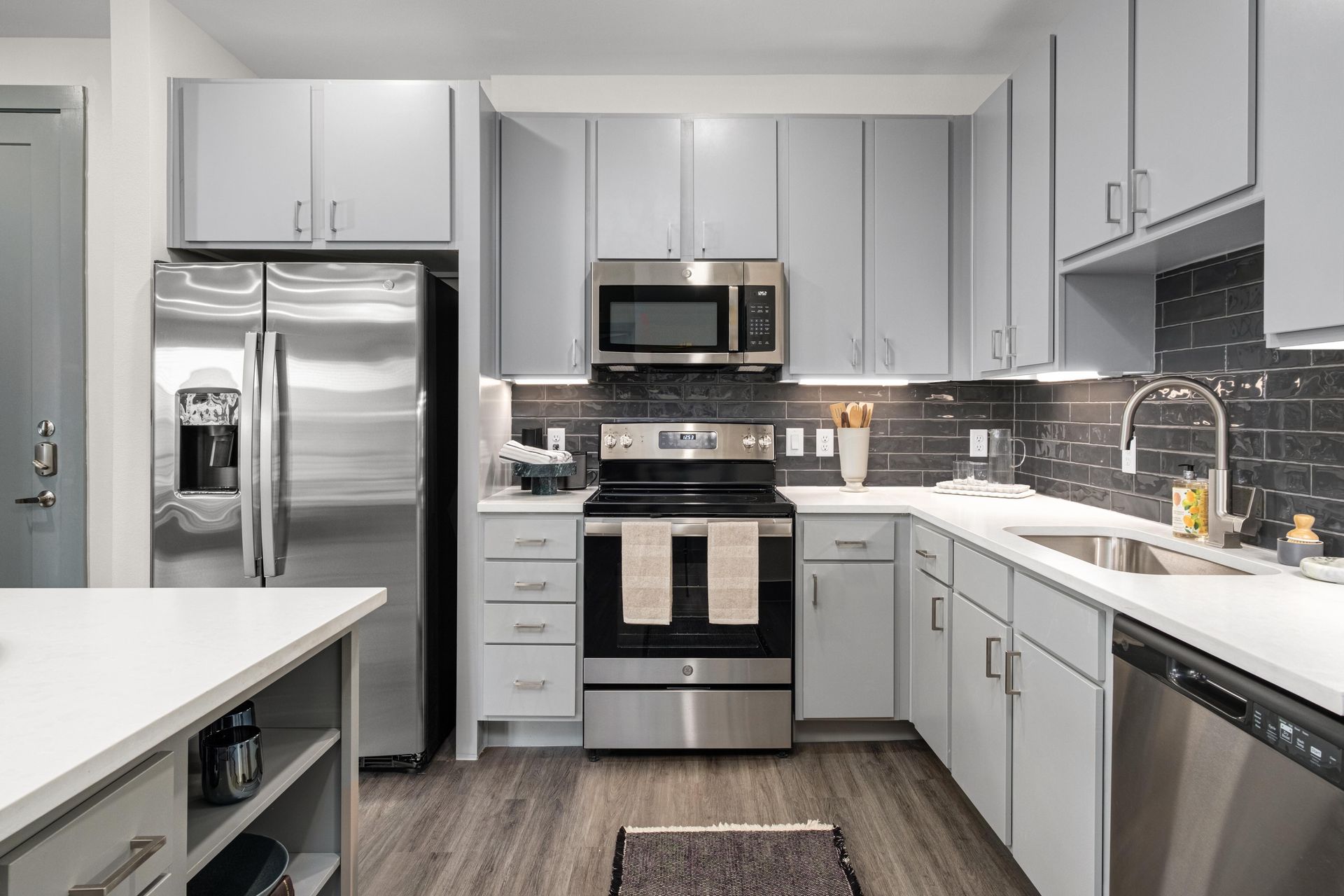 Modern apartment kitchen with light gray cabinets, white countertops, dark subway tile backsplash, wood-look flooring, and all stainless steel appliances.