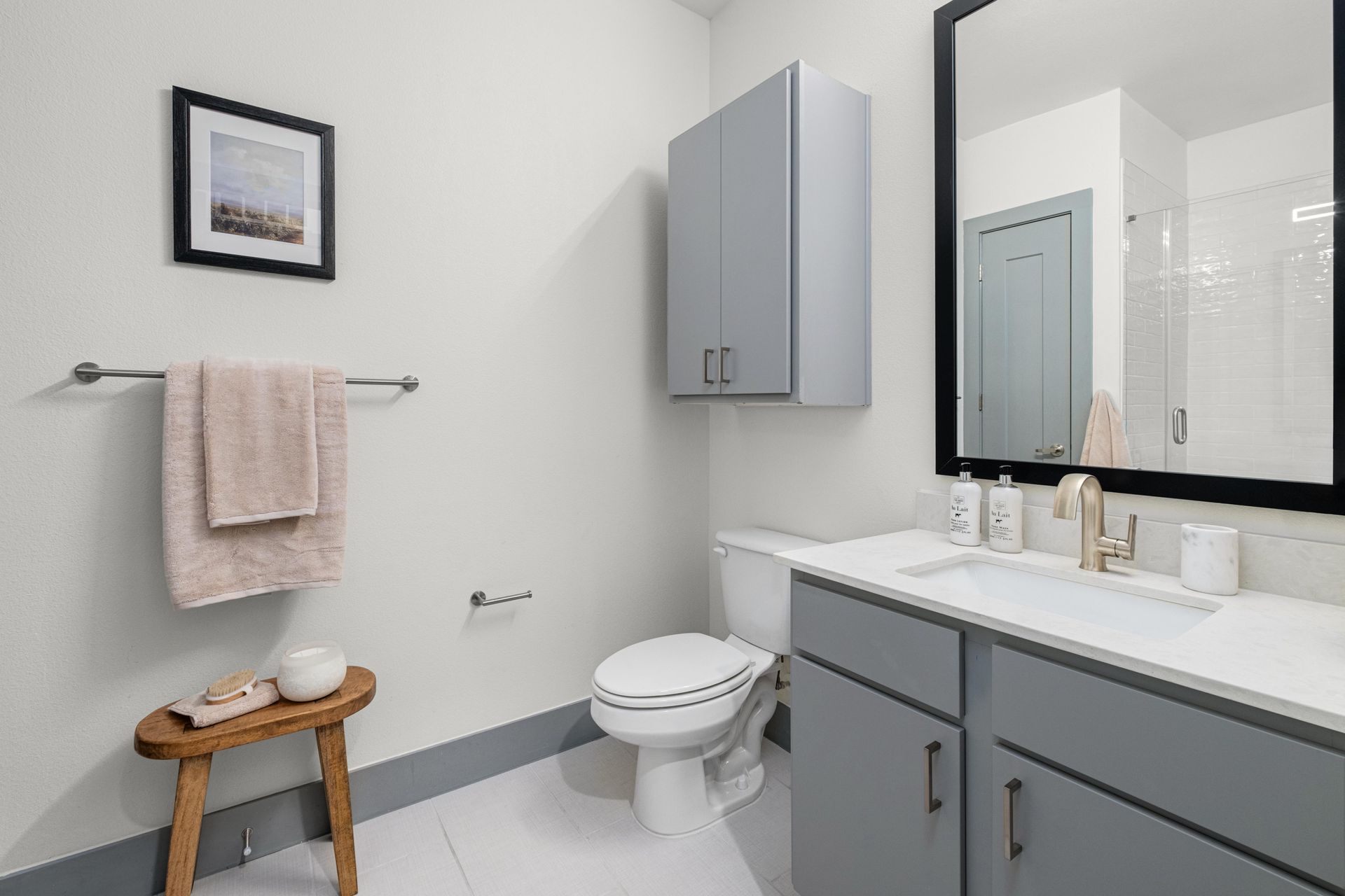Modern apartment bathroom at The Anderson has white walls, toilet, gray vanity with a sink, and a large mirror. Gray cabinets & glass shower visible.