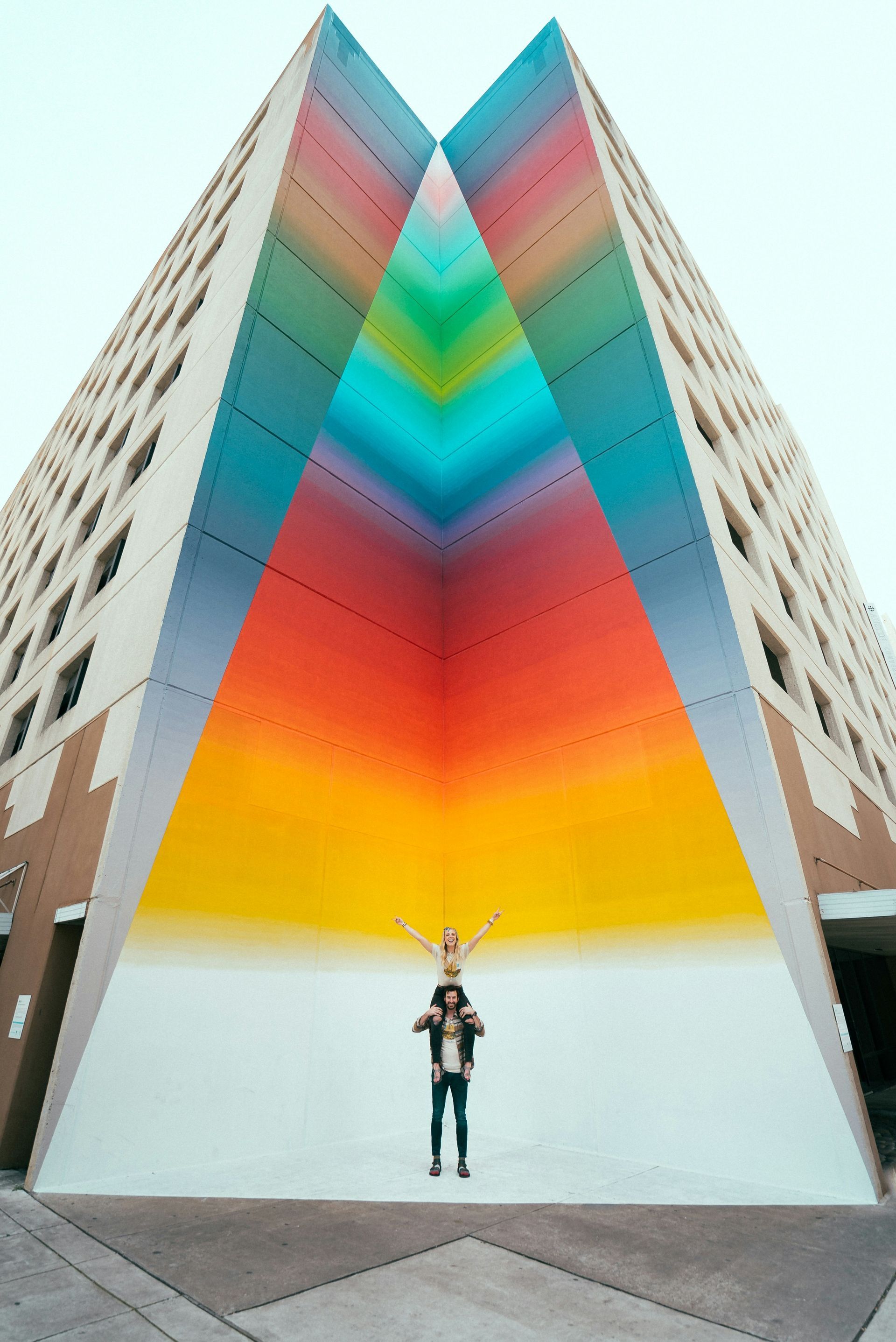 A man is carrying a child on his shoulders in front of a colorful building.