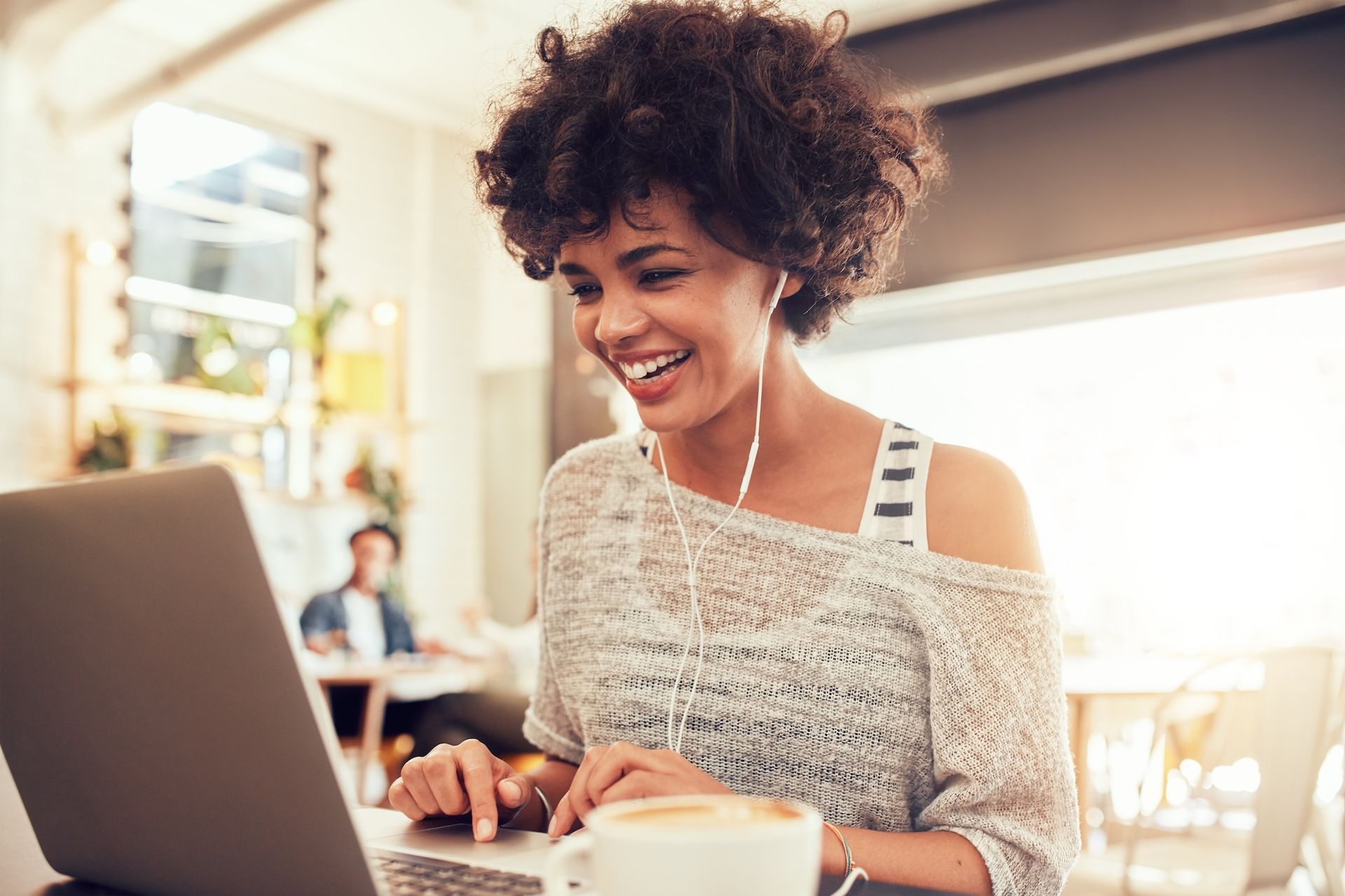 Woman smiles while using a laptop, wearing earphones. Sitting at a cafe table with a coffee.