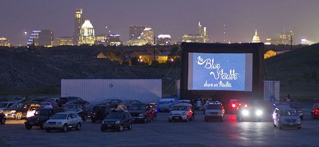 A nighttime view of a drive-in movie theater, with cars parked facing a screen featuring a logo with the words
