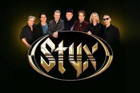 Styx band members posing behind their gold logo on a black background. They're smiling and wearing dark clothing.