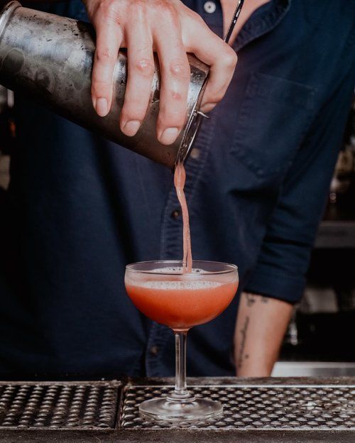 Bartender pouring a pink cocktail from a shaker into a coupe glass. Dark blue shirt and bar setting.