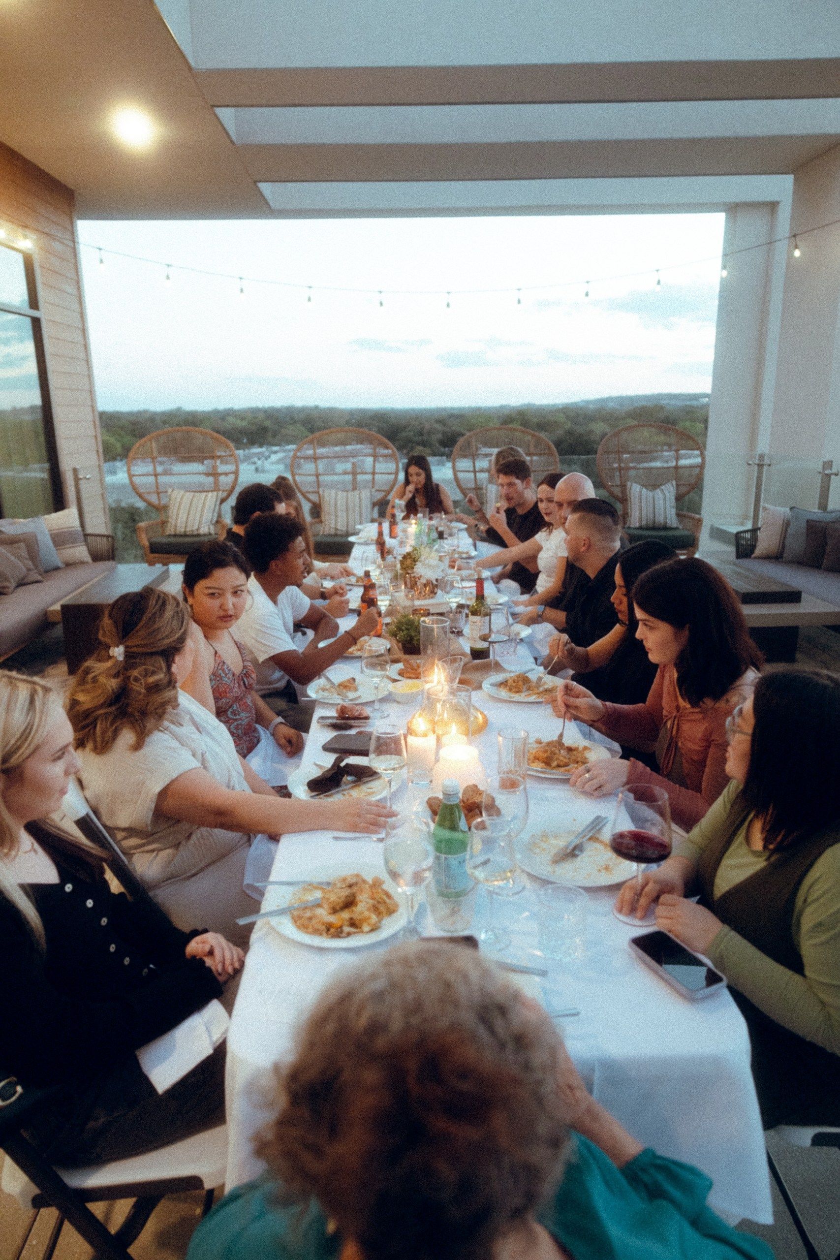 People seated at a long table, dining outdoors at dusk. Candles and food adorn the table.